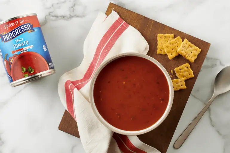 A bowl of Progresso Spicy Tomato Soup next to a napkin, a spoon, and some crackers