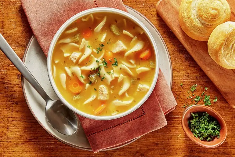 Bowl of Chicken and Homestyle Noodle soup with napkin, spoon, roll, and parsley flakes on wooden table